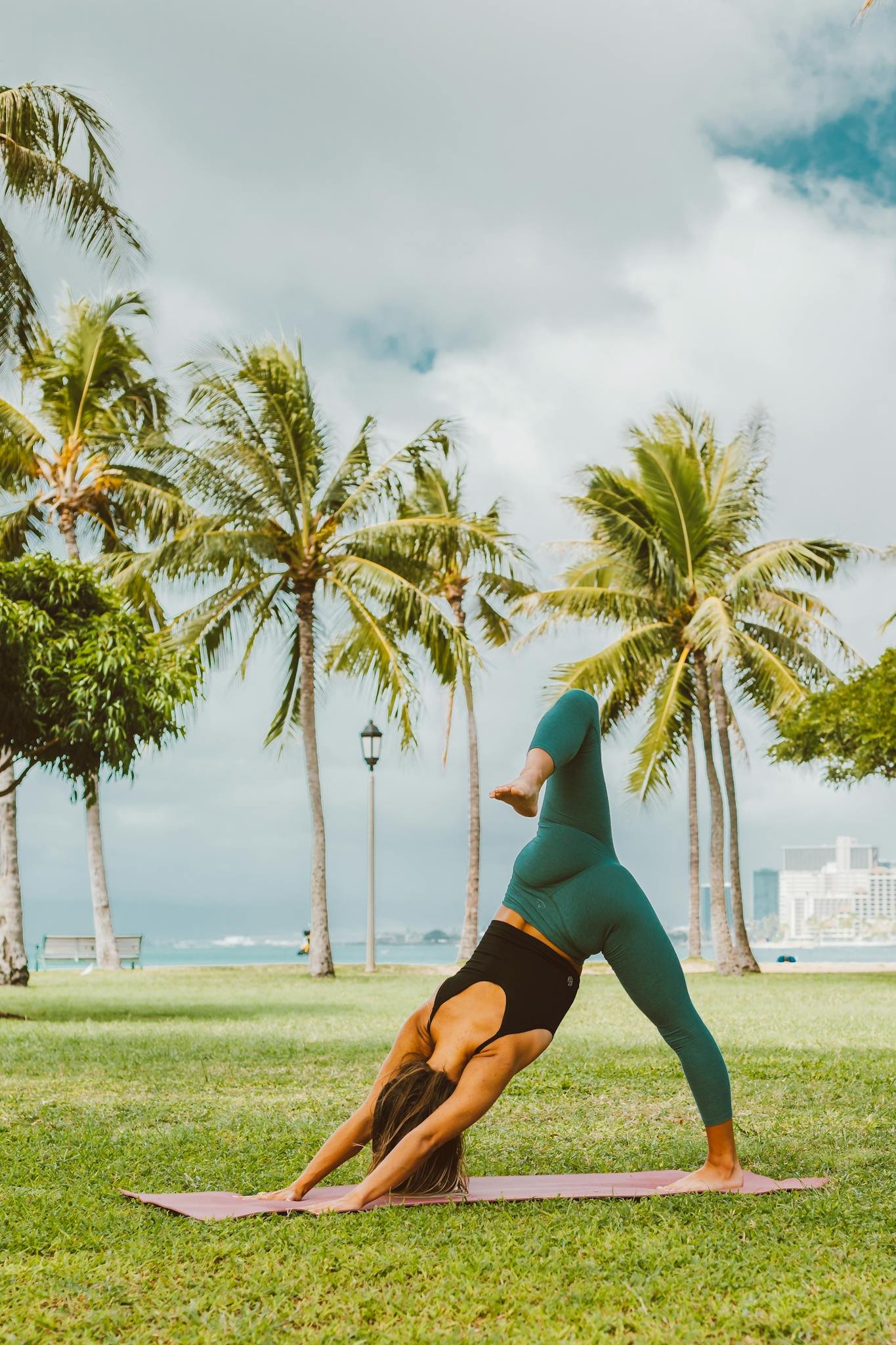 Woman practicing yoga outdoors on a sunny day with palm trees and cityscape in the background.